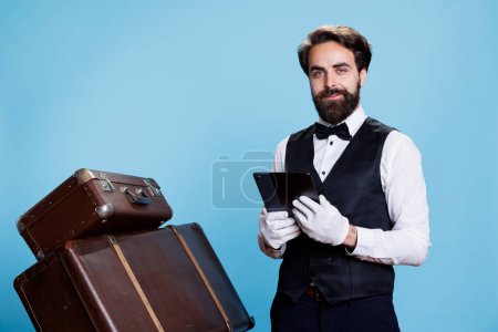 Photo for Hotel concierge checks online reservations on gadget, standing next to suitcases for safekeeping. Bellhop showcases his commitment in providing excellent services, important role. - Royalty Free Image