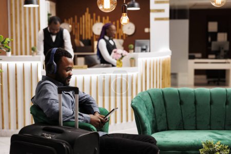 Téléchargez les photos : Jeune voyageur afro-américain souriant attendant une chambre à l'hôtel. Homme noir dans les écouteurs en utilisant une tablette numérique tout en étant assis dans le hall avec des bagages, regarder un film, attendre le processus d'enregistrement - en image libre de droit