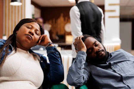 Téléchargez les photos : Jeune couple afro-américain fatigué faisant la sieste dans le hall de l'hôtel, arrivant trop tôt à la station, se reposant après un long vol. Touristes épuisés dormant en voyage. Concept des personnes et du décalage horaire - en image libre de droit