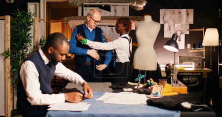 Antiguo cliente vistiendo chaqueta de traje en taller de sastrería, ropa adecuada con medidas de cuerpo hechas a medida. Cliente masculino colaborando con el equipo de couturiers en el taller de moda.