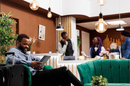 Téléchargez les photos : Heureux homme afro-américain détendu touristique tenant tablette numérique envoyer un message à la famille à l'arrivée à l'hôtel. sourire noir guy voyageur assis avec bagages dans lobby enregistrement en ligne - en image libre de droit