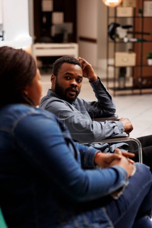 Photo for Young African American guy wheelchair user traveling with companion, disabled man tourist sitting in hotel lobby talking with girlfriend or wife while waiting for check-in, traveler with disabilities - Royalty Free Image