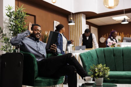 Téléchargez les photos : Joyeux jeune voyageur afro-américain souriant assis dans le hall de l'hôtel ayant un appel vidéo en attendant l'enregistrement. Touriste tenant tablette numérique, saluant la main ami regardant appareil photo - en image libre de droit