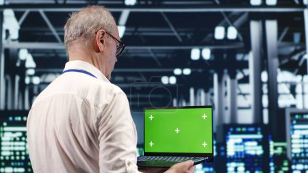 Capable IT professional navigating network of server clusters in industrial hub. Expert with green screen laptop ensuring proper operations, doing optimizations and maintenance in data center