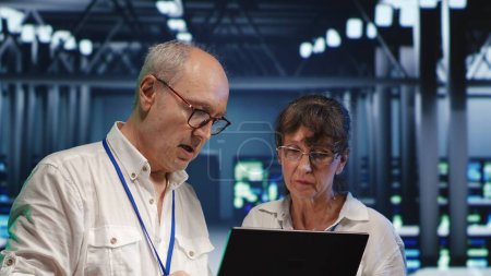 Photo for Efficient technical support executives looking around server hub, using laptop to crosscheck disaster recovery plan and assess mainframes in need of replacement, preventing hazards - Royalty Free Image