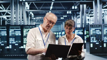 Elderly employees monitoring green energy data center infrastructure. Server systems using renewable sources employed to minimize consumption and reduce cloud computing business carbon footprint