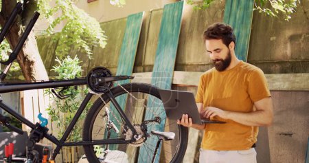 Sports-loving healthy man using laptop for enhancing safety of bike components for leisure cycling. Active male cyclist holding minicomputer for instructions to help repair damaged bicycle wheel.