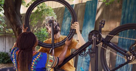 Engagierter Freund und Freundin bei der jährlichen Wartung von Fahrradreifen für sicheres Radfahren im heimischen Garten. Jugendliches multiethnisches Paar montiert Rad erfolgreich auf modernem Fahrradrahmen.