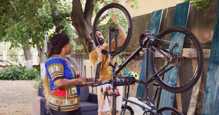 Mujer afroamericana en busca de minicomputador para ayudar a los jóvenes a reparar bicicleta dañada. Pareja amante de los deportes que fija la cadena de bicicleta mientras usa el ordenador portátil para el ciclismo al aire libre anual de verano.