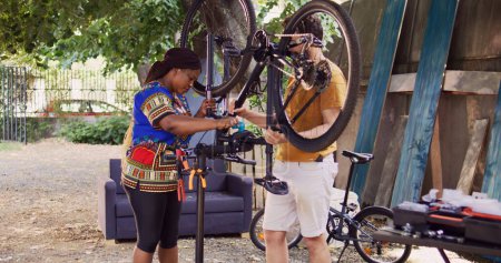 Youthful multiracial couple repairing damaged bike for summer leisure cycling. Athletic black woman helping caucasian man by clamping bicycle frame to repair-stand for easy repairing outdoor.