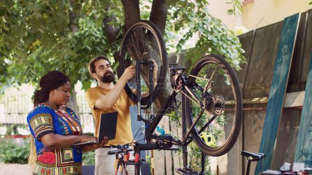 Healthy black woman searching on minicomputer to assist young man repair damaged bicycle. Boyfriend and girlfriend maintaining bike wheel while utilizing laptop for annual summer outdoor cycling.