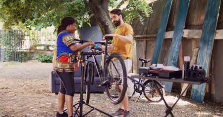 Sporty caucasian guy carrying bike and african american lady attaching frame to specialized repair-stand. Mixed-race couple secures bicycle body for thorough inspection and repair outdoor.