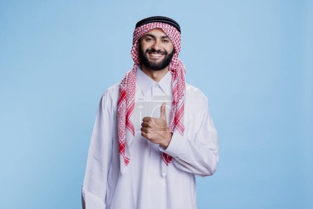 Homme arabe portant des vêtements traditionnels islamiques posant avec le pouce levé signifiant approbation et portrait d'accord. Sourire musulman dans le thobe et le ghutra montrant comme geste