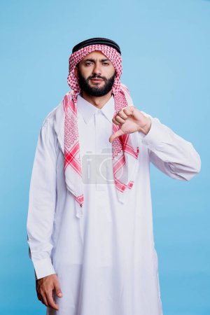 Muslim man in white thobe and checkered headscarf posing with thumb down and looking at camera with confident expression. Serious arab signifying disagreement studio portrait