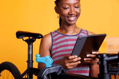 Femme noire énergique inspecte et répare soigneusement le cadre de vélo à l'aide d'un support de réparation et d'un appareil numérique. Image montrant en gros plan une cycliste tenant une tablette intelligente lors de l'entretien du vélo.