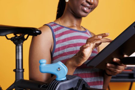 Téléchargez les photos : Vue détaillée de jeune femme noire à la recherche d'entretien de vélo sur sa tablette de téléphone. Gros plan d'une cycliste afro-américaine portant un appareil intelligent lors de l'examen de son vélo. - en image libre de droit