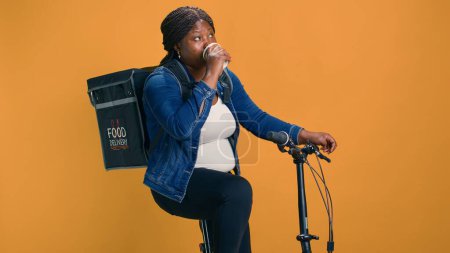 Der junge Kurier auf dem Fahrrad nimmt sich einen entspannten Moment, um Kaffee zu trinken und Pakete auszuliefern. Afroamerikanerin stillt ihren Durst mit Getränken, während sie einen effizienten Essenslieferservice anbietet.