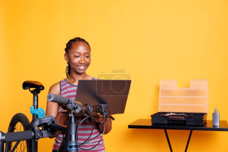 Mujer ciclista afroamericana repara bicicleta rota utilizando la caja de herramientas y el ordenador personal para buscar soluciones. Joven mujer negra trabajando en bicicleta moderna mediante el uso de minicomputador y herramientas expertas.