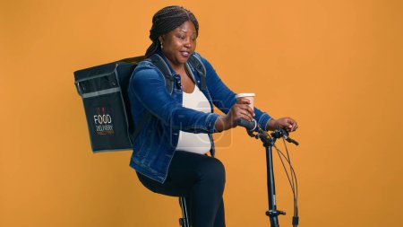 Téléchargez les photos : Livraison afro-américaine femme multitâche portant du café à la main et à vélo dans le quartier. Courrier actif en déplacement ayant un rafraîchissement énergisant rapide avant d'aller à la livraison de nourriture. - en image libre de droit