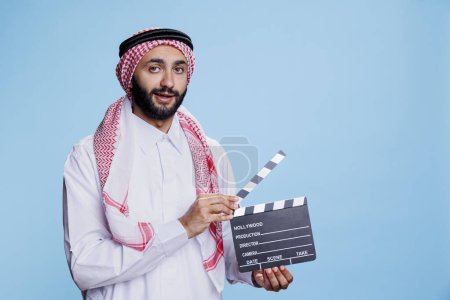 Photo for Muslim man dressed in thobe and headscarf clapping movie slate, showing film scene action studio portrait. Arab person wearing traditional clothes holding clapperboard and looking at camera - Royalty Free Image