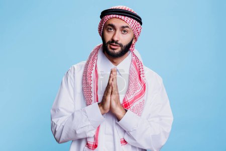 Photo for Man wearing traditional muslim thobe and ghutra clothes praying studio portrait. Arab person standing with folded hands showing symbol of faith and spirituality while looking at camera - Royalty Free Image