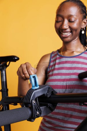 Detaillierte Ansicht einer afrikanisch-amerikanischen Frau, die das Fahrrad untersucht, anpasst und fixiert, um die Sicherheit zu gewährleisten. Das Bild zeigt eine schwarze Frau, die ein kaputtes Fahrrad an einem Reparaturstand festhält und befestigt. Nahaufnahme.