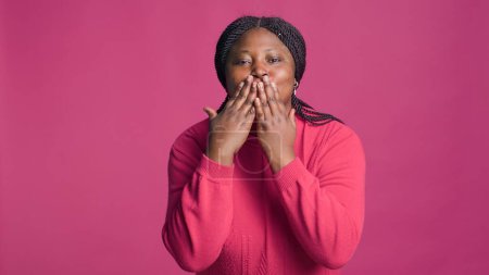 Lady with african american ethnicity blowing a kiss towards camera being lovely isolated on pink background. Stunning joyful black woman expressing love with cute kissing hand gestures.