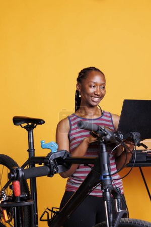 Foto de Joven afroamericana reparaciones de bicicletas dañadas utilizando el kit de herramientas y el ordenador inalámbrico. Mujer negra activa y vibrante que navega por Internet para el mantenimiento de bicicletas, frente a un fondo amarillo aislado. - Imagen libre de derechos