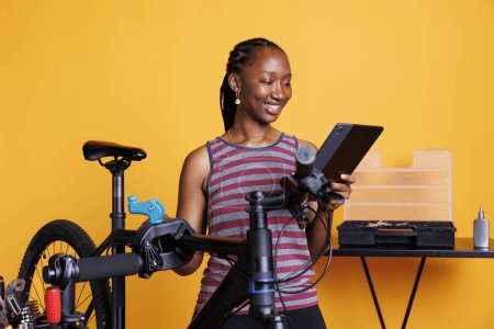 Photo for Sports-loving black woman inspecting broken bicycle, examining damaged components, researching repair solutions on tablet. African american female grasping device for bike repair online instructions. - Royalty Free Image