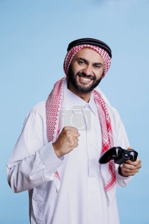 Foto de Hombre musulmán feliz mostrando gesto ganador con el puño cerrado y sosteniendo retrato inalámbrico del estudio de joystick. Árabe vestido de pie tradicional con mando de consola y videojuego ganador - Imagen libre de derechos