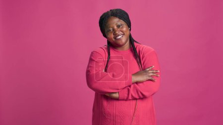 Enthusiastic black woman standing in front of a vibrant pink background with arms crossed on her chest. African american fashionista elegantly posing against isolated pink backdrop.