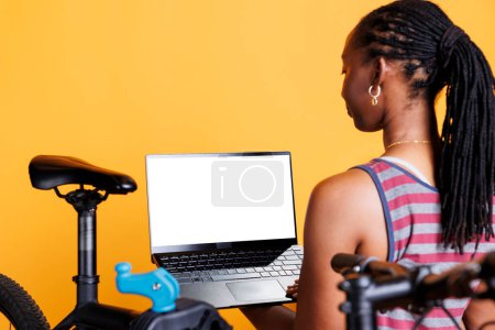 La dama afroamericana examina expertamente la bicicleta rota y usa una computadora portátil que muestra una plantilla de maqueta cromakey en blanco para orientación. Mujer joven que lleva miniordenador con una pantalla blanca.