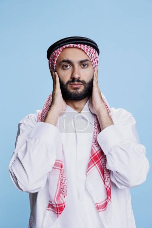Photo for Man wearing traditional muslim clothes covering ears with hands while showing hear no evil three wise monkeys concept. Arab person ignoring loud sound and looking at camera - Royalty Free Image