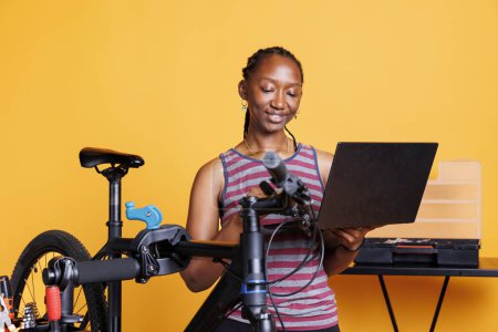 Foto de Deportiva mujer afroamericana reparando bicicleta rota con ayuda de Internet en el portátil digital. Mujer negra joven examinando y fijando componentes dañados con el juego de herramientas y el ordenador personal. - Imagen libre de derechos