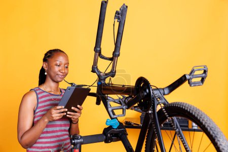 Photo for Image showing african american female holding a tablet and inspecting damaged bike components. Active sporty black woman using smart digital device to research bike repair instructions. - Royalty Free Image