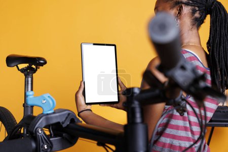 Ver en detalle de afroamericana hembra sosteniendo gadget con plantilla de cromakey espacio de copia en blanco y una bicicleta rota cercana. Mujer negra usando tableta digital con pantalla blanca maqueta para mantener la bicicleta.