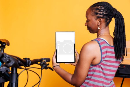 Primer plano de una mujer negra sosteniendo una tableta digital con una pantalla blanca aislada. Señora afroamericana que lleva dispositivo inteligente con una pantalla de plantilla de cromakey en blanco para la reparación de bicicletas.