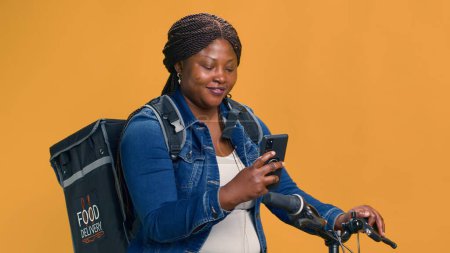 Photo for Active black woman delivering food on bicycle with cell phone in hand. African american female checking delivery app on smartphone for transportation of package. Portrait shot, smiling at camera. - Royalty Free Image