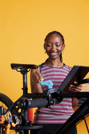 Photo for Active black woman examining damaged bicycle parts and using digital device for repair research. Sporty female multitasking, servicing bike and utilizing her digital tablet for guidance. - Royalty Free Image