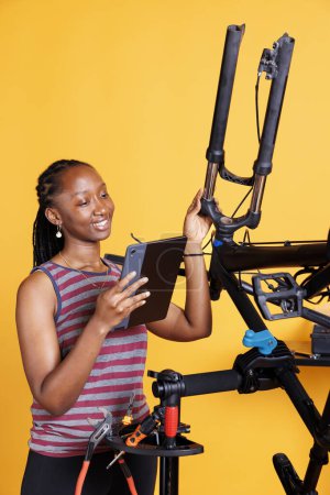 Mujer atlética negro busca instrucciones de reparación de bicicletas en la tableta digital inteligente. Sonriente señora afroamericana agarrando dispositivo e inspeccionando bicicleta para reparaciones sobre fondo amarillo.