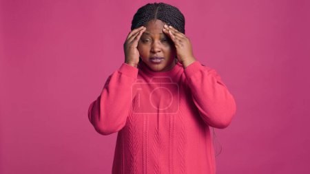 Fashionable black woman wearing pink sweater suffering a bad headache. Female african american beauty covering face in front of camera showcasing having a migraine.