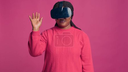 Photo for Innovative young black woman enjoys futuristic simulation with interactive device while wearing 3D virtual reality glasses in studio. African american trendy fashionista using modern VR headset. - Royalty Free Image