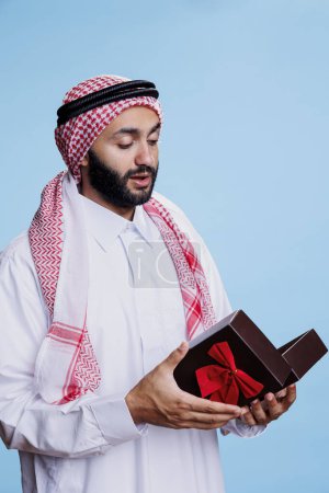 Téléchargez les photos : Homme musulman portant des vêtements traditionnels tenant boîte cadeau avec ruban papillon et cadeau de fête d'ouverture. Arabe habillé en thobe blanc debout avec boîte cadeau en studio sur fond bleu - en image libre de droit