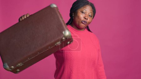 Excited female modern traveler carrying brown suitcase ready to travel in style. Fashionable african american woman in stylish outifit holding her travel carry-on in front of pink backdrop.