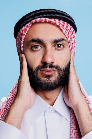 Mann mit muslimischem kariertem Kopftuch, das Ohren mit Armen bedeckt und in Großaufnahme in die Kamera blickt. Araber in traditioneller islamischer Kleidung zeigen, dass sie kein böses Drei-Affen-Konzept hören