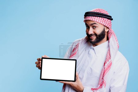 Foto de Hombre alegre musulmán que ofrece tableta digital con pantalla vacía maqueta, retrato de productos publicitarios. Sonriente persona árabe sosteniendo la pantalla táctil blanca en modo horizontal y mirando a la cámara - Imagen libre de derechos