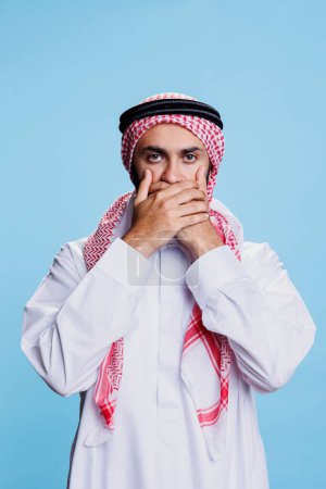 Araber in traditioneller islamischer Kleidung mit Handflächen vor dem Mund und der Bitte, ruhig zu bleiben. Muslimische Person zeigt kein böses Drei-Affen-Konzept und schaut in die Kamera