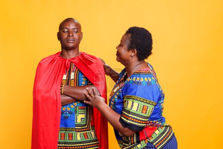 Photo for Happy wife appreciating husband in superhero cape, showing love and support. Confident african american man wearing red superman cloak standing near woman on studio background - Royalty Free Image
