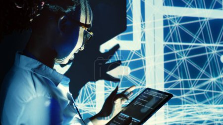 Capable supervisor overseeing dimly lit server hub, running code on tablet, doing checkup on technologically advanced machines clusters made of servers, networking systems and storage arrays