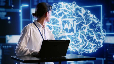 Téléchargez les photos : Admin en utilisant un ordinateur portable pour visualiser les réseaux neuronaux d'intelligence artificielle constitués de nœuds interconnectés utilisant la technologie holographique AR. Inspecteur supervisant les systèmes d'IA traitant l'information - en image libre de droit